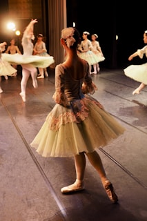 Several ballet dancers on stage wearing elegant, classical costumes are captured in motion under warm stage lighting. The dancers are dressed in pastel-colored tutus and are engaged in a graceful performance. One dancer in the foreground is standing in a poised position, while others in the background are captured mid-spin.