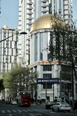 A cityscape featuring a tall building with a distinctive golden dome. The structure is surrounded by other modern high-rise buildings. There is a street scene at the foreground with cars and a red double-decker bus driving along the road. Trees and streetlights line the avenue, and pedestrians can be seen on the sidewalks. The signage on the buildings includes both Chinese and English characters.