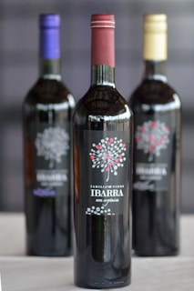 Three wine bottles are arranged with labels featuring a decorative design and the text 'Familia de Vinos Ibarra' and 'una opción'. The focus is on the front bottle with a reddish cap, while the two bottles with gold and purple caps are blurred in the background.
