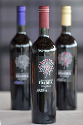 Three wine bottles are arranged with labels featuring a decorative design and the text 'Familia de Vinos Ibarra' and 'una opci&oacute;n'. The focus is on the front bottle with a reddish cap, while the two bottles with gold and purple caps are blurred in the background.