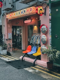 A quaint vintage shop exterior with a pink facade displays a colorful sign that reads 'Vintage Wknd'. Below the sign are three mirrors with red frames, reflecting the street. Three vibrant chairs in red, blue, and yellow are arranged outside next to potted plants. The interior appears to showcase vintage clothing on racks.