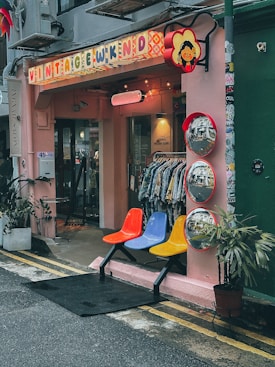 A quaint vintage shop exterior with a pink facade displays a colorful sign that reads 'Vintage Wknd'. Below the sign are three mirrors with red frames, reflecting the street. Three vibrant chairs in red, blue, and yellow are arranged outside next to potted plants. The interior appears to showcase vintage clothing on racks.
