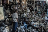 A cluttered marketplace stall filled with a wide variety of vintage and antique items. There are numerous objects including paintings, clocks, lamps, pots, and trinkets all densely packed together. A person with long hair stands amidst the chaos, seemingly examining the items closely.