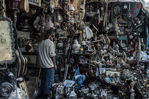 A cluttered marketplace stall filled with a wide variety of vintage and antique items. There are numerous objects including paintings, clocks, lamps, pots, and trinkets all densely packed together. A person with long hair stands amidst the chaos, seemingly examining the items closely.