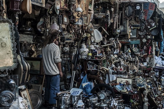 A cluttered marketplace stall filled with a wide variety of vintage and antique items. There are numerous objects including paintings, clocks, lamps, pots, and trinkets all densely packed together. A person with long hair stands amidst the chaos, seemingly examining the items closely.