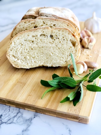 Slices of freshly baked bread are placed on a wooden cutting board. Nearby, sprigs of sage and cloves of garlic add a rustic touch. The background features a marble surface, contributing to a clean and elegant presentation.