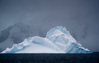 A large iceberg floats majestically in dark, icy waters under an overcast sky. The iceberg's surface is rugged and textured, with a striking blend of white and blue hues. In the background, dark, snow-covered mountains loom, partially obscured by clouds.