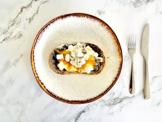 A plate with a slice of toast topped with marshmallows, bananas, and a drizzle of honey, placed on a marble surface with a knife and fork beside it.
