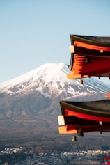 A majestic snow-capped mountain dominates the background as traditional Japanese architectural elements, with vivid red and dark brown hues, accentuate the foreground. Below, a sprawling cityscape lies at the foot of the mountain, blending into the natural surroundings.