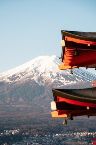 A majestic snow-capped mountain dominates the background as traditional Japanese architectural elements, with vivid red and dark brown hues, accentuate the foreground. Below, a sprawling cityscape lies at the foot of the mountain, blending into the natural surroundings.