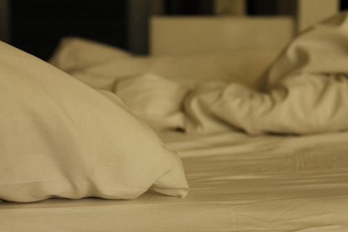 A set of wrinkled and disorderly white bed sheets and pillows on a bed, suggesting it has been used recently. The lighting is dim, giving the scene a soft and quiet atmosphere.
