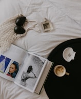 A stylish flat lay composition sits on a white fabric surface featuring two avocados in a net bag, an open magazine showing elegant photographs, a bottle of perfume, a black circular tray with a cup of coffee, and a shell-shaped dish holding a ring. The arrangement exudes an artful balance of fashion and simplicity.