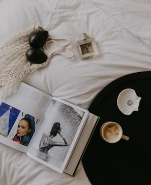A stylish flat lay composition sits on a white fabric surface featuring two avocados in a net bag, an open magazine showing elegant photographs, a bottle of perfume, a black circular tray with a cup of coffee, and a shell-shaped dish holding a ring. The arrangement exudes an artful balance of fashion and simplicity.