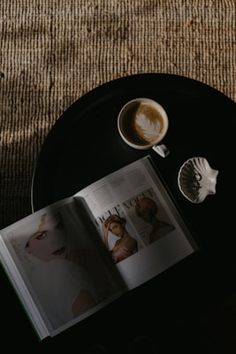 A top-down view of a black round table featuring an open fashion magazine displaying images of women, a cup of coffee with latte art, and a ceramic seashell on the side. The table is placed on a textured rug with earthy tones.