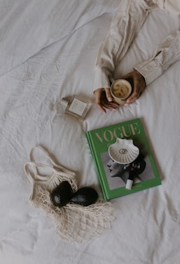 A cozy composition on a white bed featuring a person holding a cup of frothy coffee. Nearby lie a bottle of perfume, a green Vogue magazine adorned with a shell and a lipstick, and a net bag containing two avocados.