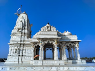A beautifully ornate white marble temple with intricate carvings and architectural details stands against a clear blue sky. The structure features multiple domes and spires, with decorative sculptures and pillars showcasing detailed craftsmanship. There is a flagpole with a flag atop one of the spires, adding to the temple's grandeur.