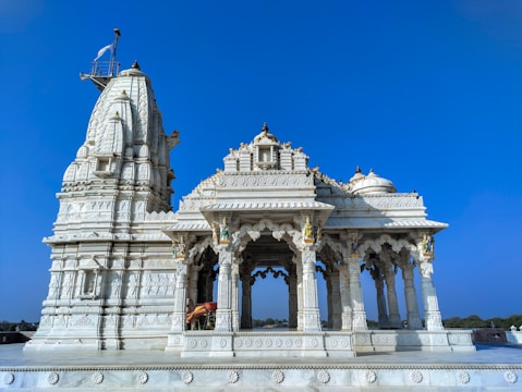 A beautifully ornate white marble temple with intricate carvings and architectural details stands against a clear blue sky. The structure features multiple domes and spires, with decorative sculptures and pillars showcasing detailed craftsmanship. There is a flagpole with a flag atop one of the spires, adding to the temple's grandeur.