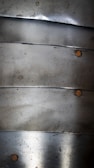 Several overlapping metal plates with a weathered texture are arranged vertically. Each plate is fastened by small, round rivets. The surface shows signs of oxidation and minor scratches.