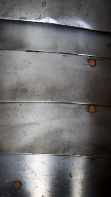 Several overlapping metal plates with a weathered texture are arranged vertically. Each plate is fastened by small, round rivets. The surface shows signs of oxidation and minor scratches.