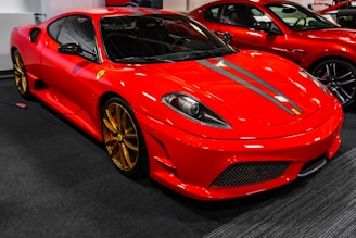 A red sports car with sleek lines and golden rims is prominently displayed in a well-lit showroom. The car has a low profile, aerodynamic design, and a distinctive hood with gray racing stripes. Another similar sports car is partially visible in the background.