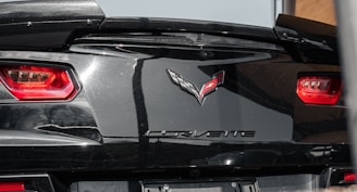 Close-up view of the rear end of a black sports car showing its red tail lights and the Corvette emblem prominently displayed on the trunk.