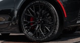 A close-up view of a sleek black car wheel with intricate alloy rims and visible tread on the tire. The wheel features red brake calipers that add a striking contrast against the dark elements. The car's bodywork reflects a shiny surface, hinting at a well-polished, high-performance vehicle.