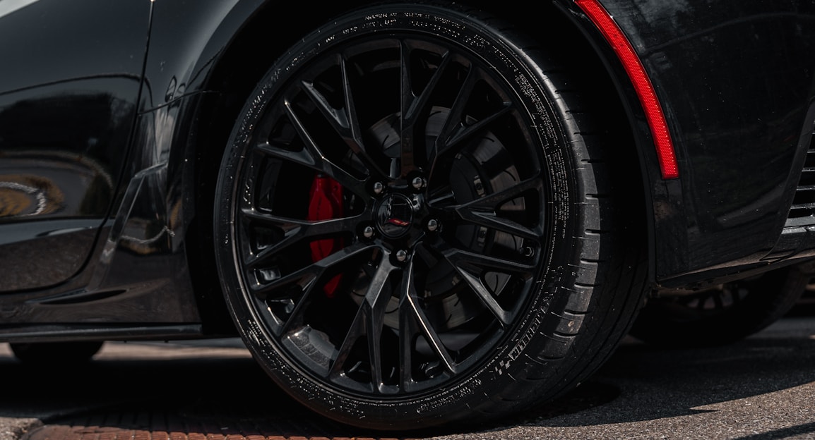A close-up view of a sleek black car wheel with intricate alloy rims and visible tread on the tire. The wheel features red brake calipers that add a striking contrast against the dark elements. The car's bodywork reflects a shiny surface, hinting at a well-polished, high-performance vehicle.