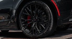 A close-up view of a sleek black car wheel with intricate alloy rims and visible tread on the tire. The wheel features red brake calipers that add a striking contrast against the dark elements. The car's bodywork reflects a shiny surface, hinting at a well-polished, high-performance vehicle.