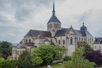 A large, historic stone church with multiple sections, arches, and a tall central spire. Surrounding the church are lush green trees and bushes, adding vibrancy to the scene. The sky is overcast, contributing to a serene and somewhat somber atmosphere.