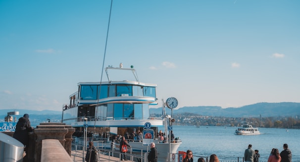 A docked boat with large windows and modern design sits by the waterfront. People are gathered around the dock, some walking and others appearing to be waiting. A clock is mounted on a pole next to the dock. Another boat can be seen in the water, and the background reveals a hilly landscape under a clear blue sky.