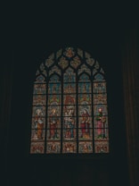 A detailed stained glass window featuring religious figures and intricate designs. The window is arched and comprises multiple panels with vibrant colors, including depictions of people in robes and ornate patterns. The setting suggests a church or cathedral environment, highlighting craftsmanship and historical artistry.