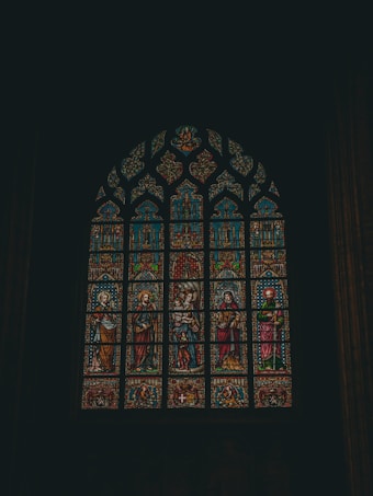 A detailed stained glass window featuring religious figures and intricate designs. The window is arched and comprises multiple panels with vibrant colors, including depictions of people in robes and ornate patterns. The setting suggests a church or cathedral environment, highlighting craftsmanship and historical artistry.