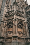 An ornate Gothic architectural detail with intricate stone carvings and sculptures adorning a large corner section of a historic building. The structure features pointed arches, elaborate tracery, and detailed figurative elements. Scaffolding is present on the right side, indicating ongoing restoration work.