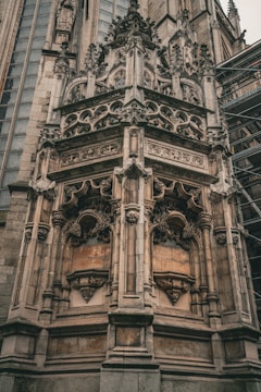 An ornate Gothic architectural detail with intricate stone carvings and sculptures adorning a large corner section of a historic building. The structure features pointed arches, elaborate tracery, and detailed figurative elements. Scaffolding is present on the right side, indicating ongoing restoration work.