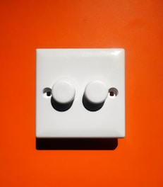 A white light switch panel with two round dials is mounted against a vibrant orange wall.