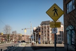 A city street scene featuring a bright yellow pedestrian crossing sign with an additional 'Seniors' sign attached below. The background reveals a mix of modern and older brick buildings, with visible traffic lights, road signs, and urban elements like street poles and trees under a clear blue sky.