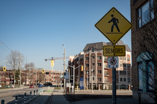 A city street scene featuring a bright yellow pedestrian crossing sign with an additional 'Seniors' sign attached below. The background reveals a mix of modern and older brick buildings, with visible traffic lights, road signs, and urban elements like street poles and trees under a clear blue sky.