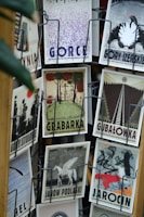A collection of vintage-style postcards displayed on a rotating rack, featuring artistic illustrations and typography. Each postcard has a unique design and depicts different themes, such as landscapes, animals, and abstract art. The cards are displayed in metal holders attached to a wooden stand.
