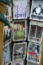 A collection of vintage-style postcards displayed on a rotating rack, featuring artistic illustrations and typography. Each postcard has a unique design and depicts different themes, such as landscapes, animals, and abstract art. The cards are displayed in metal holders attached to a wooden stand.