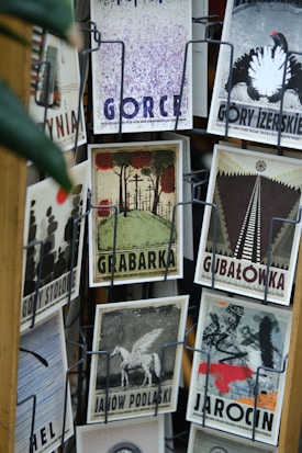 A collection of vintage-style postcards displayed on a rotating rack, featuring artistic illustrations and typography. Each postcard has a unique design and depicts different themes, such as landscapes, animals, and abstract art. The cards are displayed in metal holders attached to a wooden stand.