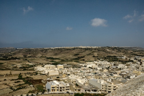A sprawling landscape with a clear, blue sky above. The view encompasses a variety of agricultural fields and a densely packed cluster of white and beige buildings in the foreground. The terrain is slightly hilly and stretches into the distance, with patches of greenery and cultivated land.