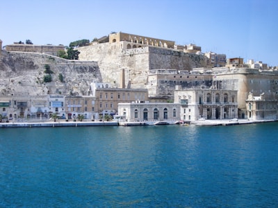 A historical seaside cityscape features majestic limestone buildings and fortifications. The structures sit prominently on elevated ground overlooking the deep blue water. Architectural elements such as arches and detailed facades highlight the region's rich heritage. Vegetation sparsely dots the scene, adding touches of green.