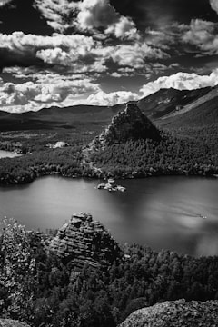 A serene landscape featuring a calm lake surrounded by dense forest and rugged rock formations. The dramatic sky filled with thick, fluffy clouds adds an element of grandeur to the natural scenery. The image is captured in black and white, highlighting contrasts and textures.