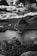 A serene landscape featuring a calm lake surrounded by dense forest and rugged rock formations. The dramatic sky filled with thick, fluffy clouds adds an element of grandeur to the natural scenery. The image is captured in black and white, highlighting contrasts and textures.