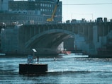 A large bridge under construction or renovation spans over a body of water. Below the bridge, there is a barge on the water with a 'DAM' sign visible. The bridge is surrounded by scaffolding and protective coverings, with a crane present on top. Buildings are visible in the background, and the water reflects the surrounding structures.