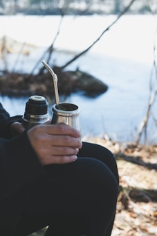 A person is sitting outdoors near a body of water, holding a gourd with a metal straw, typically used for drinking yerba mate. There is also a thermos in their hand. The environment is natural with trees and a lake in the background, suggesting a peaceful and tranquil setting.