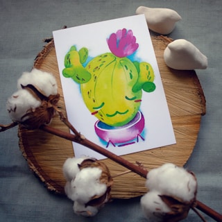 A colorful drawing of a cartoon cactus with a pink flower on top. The cactus is in a purple pot and has a cute, smiling expression. The drawing is placed on a round wooden surface, surrounded by cotton flowers and two white ceramic bird figures.