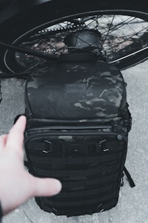 A black, camo-patterned backpack is placed against the backdrop of a bicycle wheel. A hand reaches out towards the backpack, which appears to be rugged and designed for tactical or outdoor use.