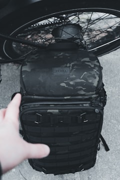 A black, camo-patterned backpack is placed against the backdrop of a bicycle wheel. A hand reaches out towards the backpack, which appears to be rugged and designed for tactical or outdoor use.