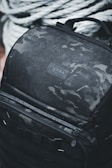 A rugged black backpack with a camouflage pattern is covered in water droplets, indicating it is water-resistant. The backpack features multiple zippers and handles, and it is positioned against a backdrop of thick, coiled ropes.